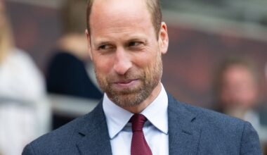 Prince William smiling while wearing a dark gray suit, a dark red tie, and a white shirt