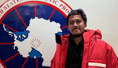 A man poses in a red parka in front of an emblem.