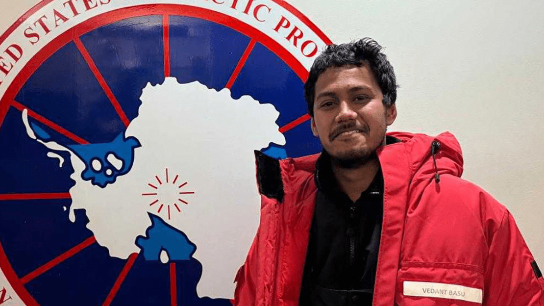 A man poses in a red parka in front of an emblem.