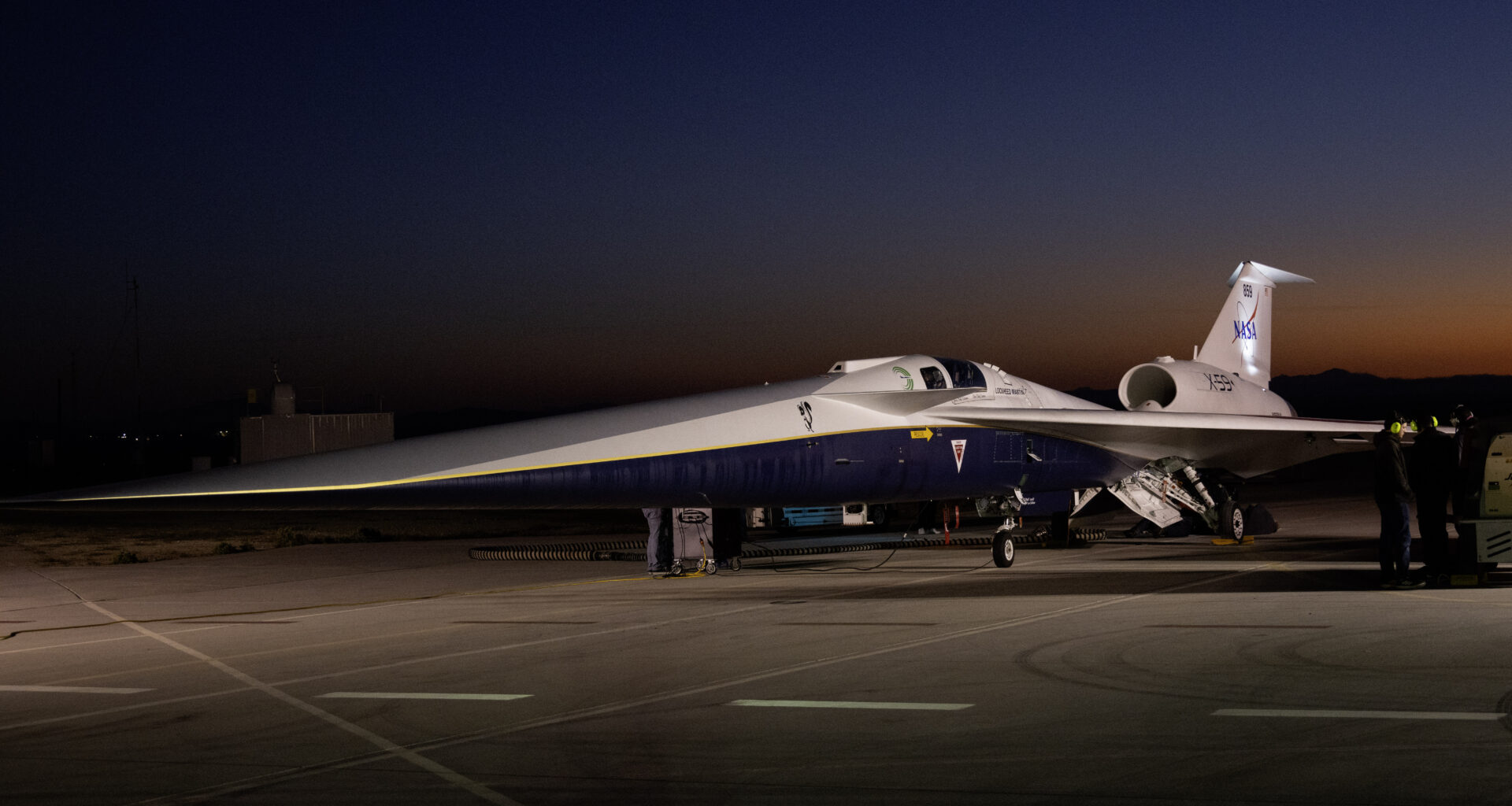 An aircraft resting on a section of runway as seen from the side. The X-59 has a long, thin nose that accounts for nearly a third of its length, along with sleek wings and an engine mounted above its body, just below its tail. The early morning sky is dark in the background with the sunrise just starting to emerge.