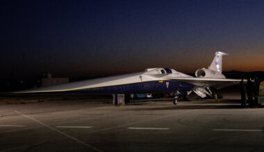 An aircraft resting on a section of runway as seen from the side. The X-59 has a long, thin nose that accounts for nearly a third of its length, along with sleek wings and an engine mounted above its body, just below its tail. The early morning sky is dark in the background with the sunrise just starting to emerge.