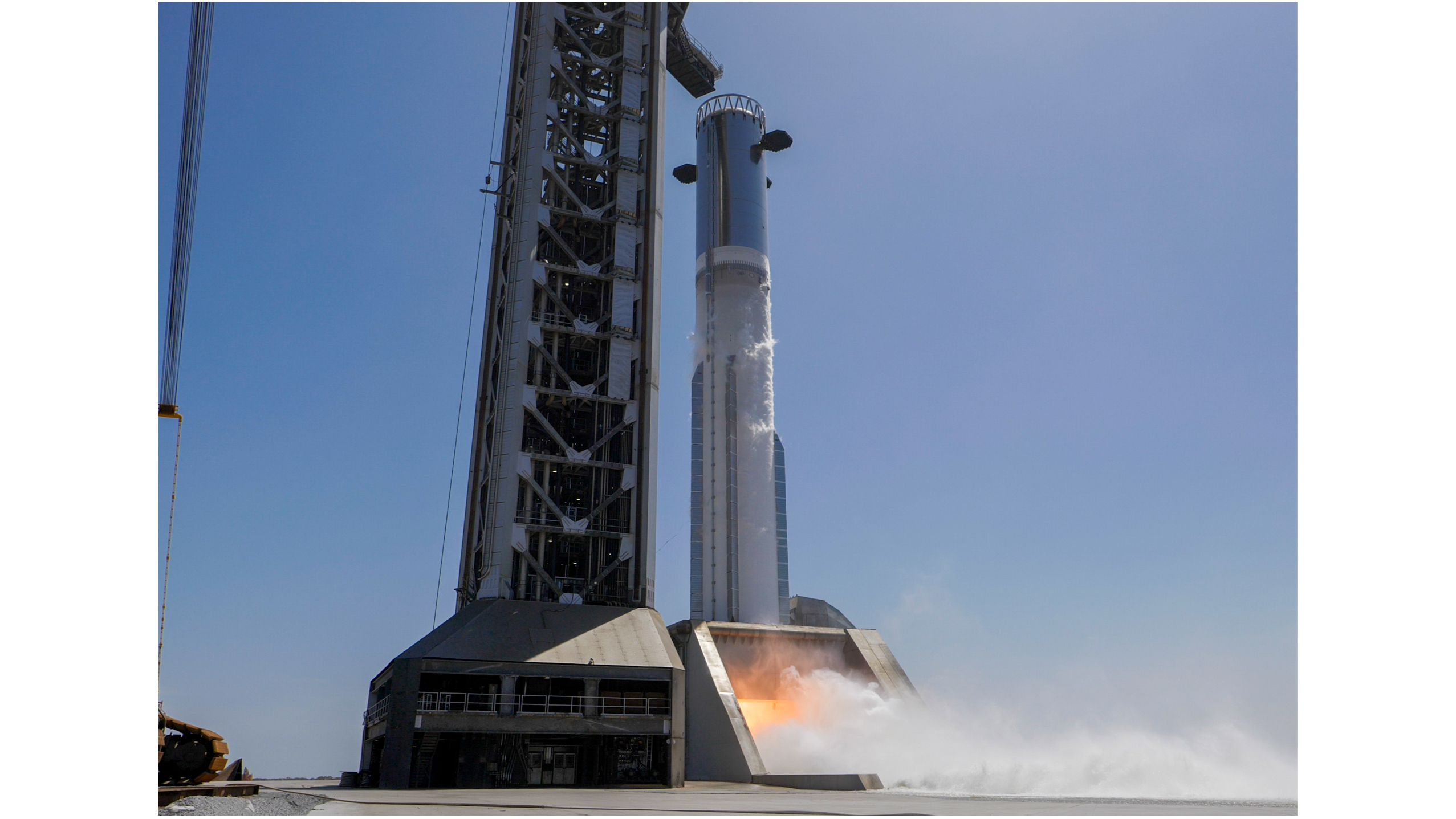ground-level view of a large silver rocket firing its engines on the launch pad beneath a blue sky