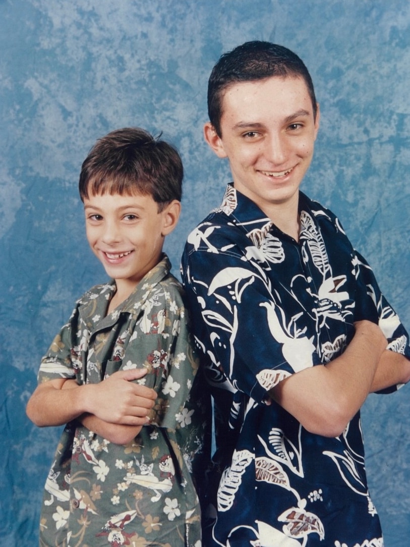 A toung boy and a young teenager stand next to each other in shot-sleeve shirts, smiling at the camera for a portrair.