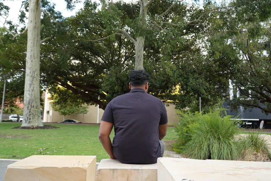 A man wearing a dark polo shirt sits on a park bench with his back turned to the camera.