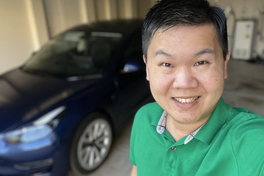 A smiling man in a green shirt taking a selfie in front of a navy car.