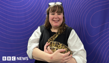 A woman, Vikki Buxton-Helyer, who is wearing glasses on top of her head and a white and black top, is holding a large yellow and black spotted snake in her arms. She is smiling and standing in front of a purple background.