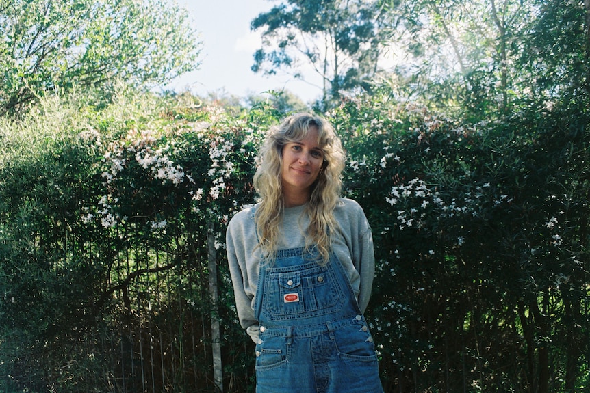 A woman wearing overalls with long curly blonde hair smiles among foliage.