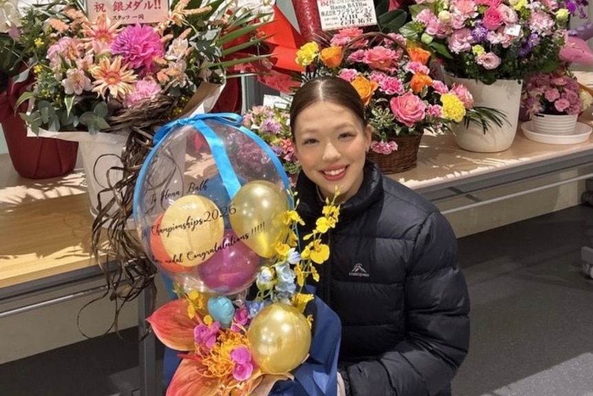 Hana Bath crouches in front of several bouquet of flowers and balloons and holds one of the bouquets