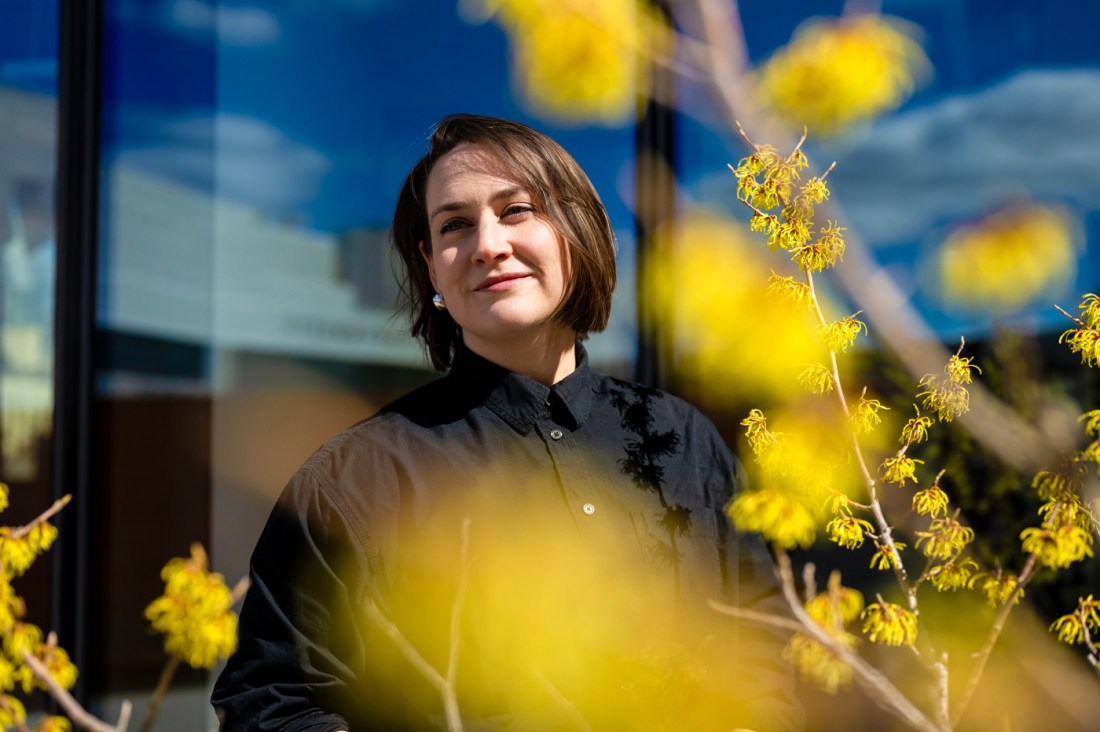 Brie Reid poses for a portrait with yellow flowers in the foreground and the reflection of sky in windows behind her.