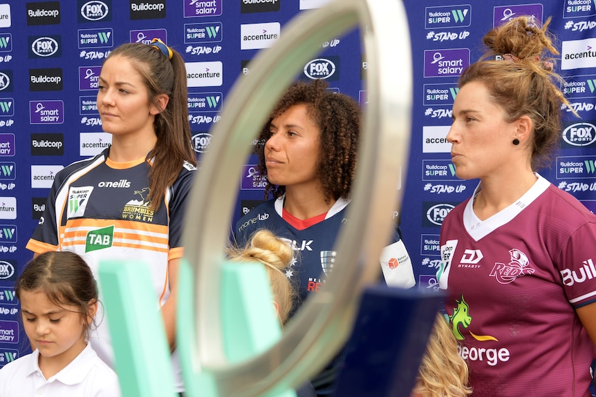 Three women pose in front of a Super W trophy with young girls in front of a banner.