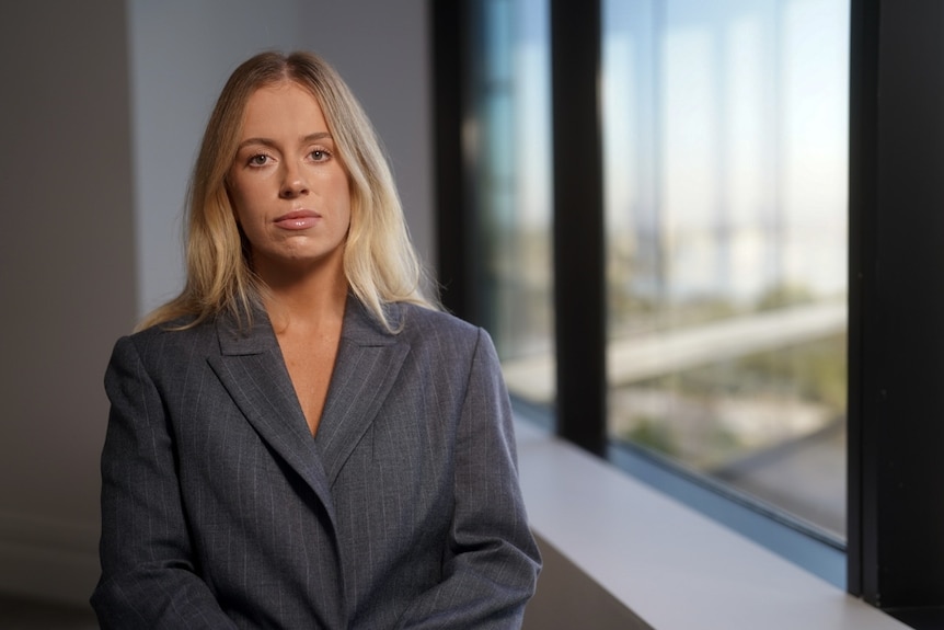 A woman in a grey jacket with blonde hair looks at the camera with a solemn look.