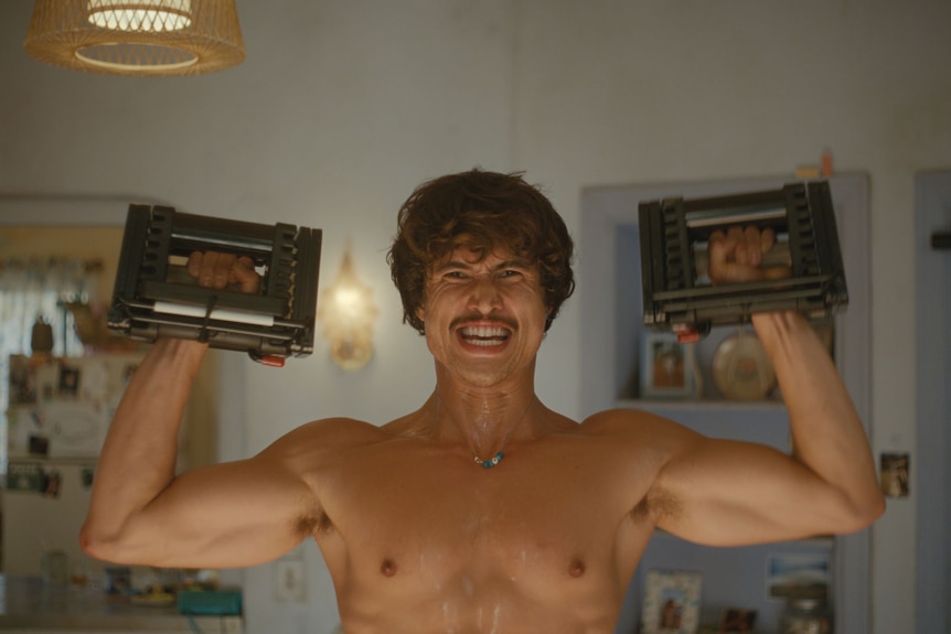 A shirtless young man, standing in his loungeroom, strains as he lifts weights above his head.