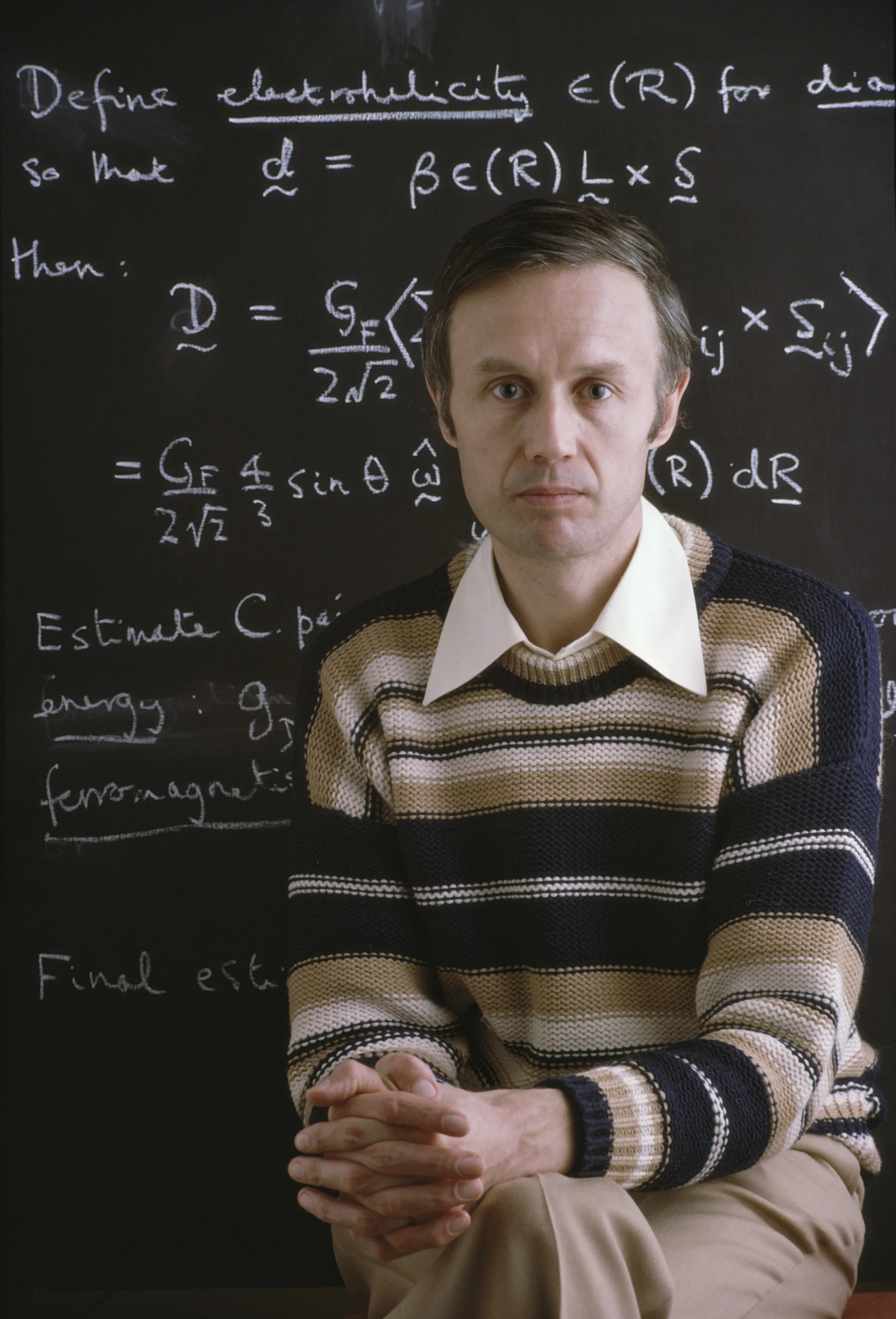 Sir Anthony James Leggett, a physics professor and Nobel Prize winner, sits in front of a chalkboard covered in physics equations.