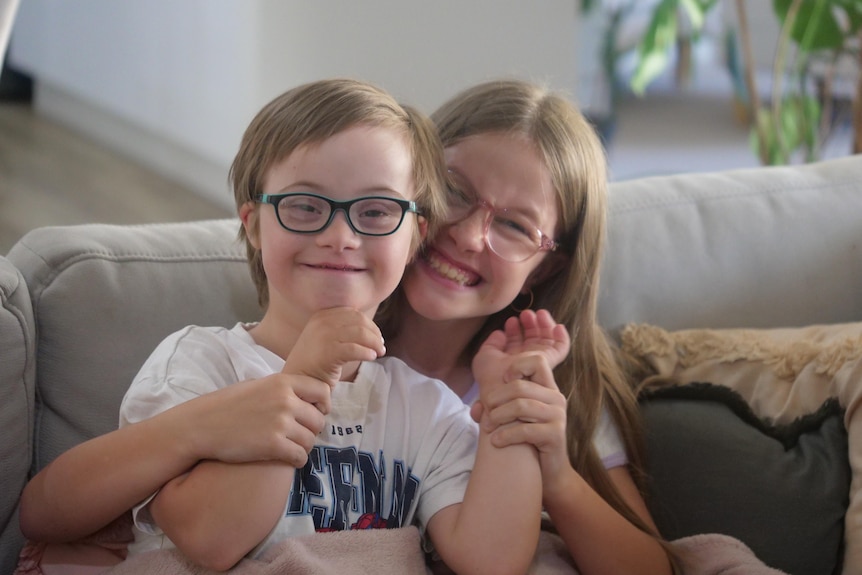 A smiling boy and girl, both with galsses and fair hair, sit together on a couch.