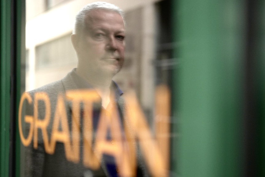 A window reflection of a man with short white hair and wearing a suit. The word "grattan" is on the window