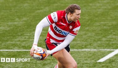 Emma Sing holds the rugby ball down to her right leg in both hands as she looks over her left shoulder while on the pitch during a match for Gloucester-Hartpury