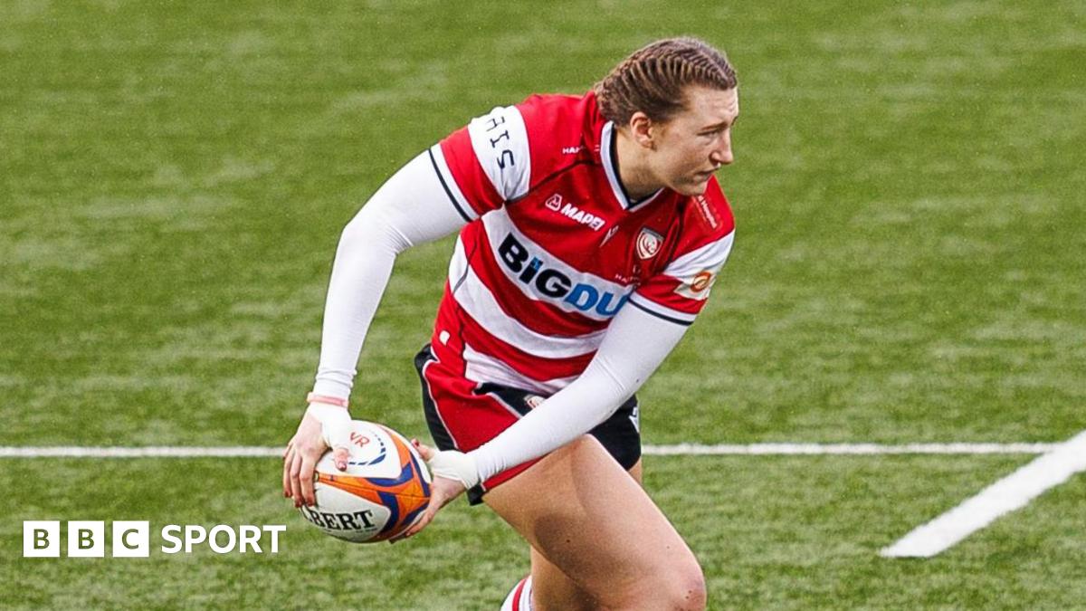 Emma Sing holds the rugby ball down to her right leg in both hands as she looks over her left shoulder while on the pitch during a match for Gloucester-Hartpury