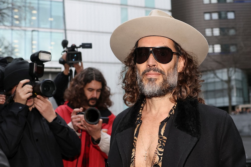 Russell Brand wears sunglasses and a white cowboy hat outside court as his photo is taken by media