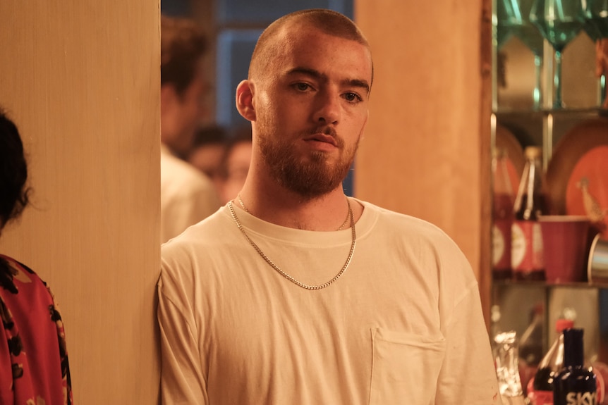 Angus Cloud in character as Fezco wearing a white t-shirt and gold chain, leaning on a wall