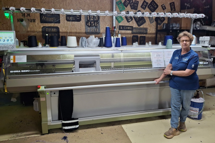 Older woman standing in front of a large grey metal knitting machine. She is wearing blue jeans and blue shirt