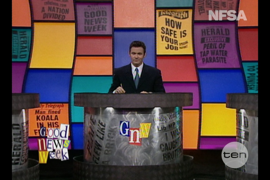 A TV still of Paul McDermott, mid-30s, seated behind a desk on a TV set, news headlines on the bright wall behind him.