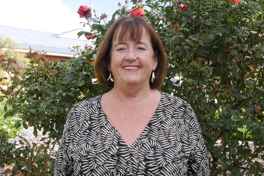 A woman with shoulder length brown hair stands in front of rose bushes, smiling at the camera.
