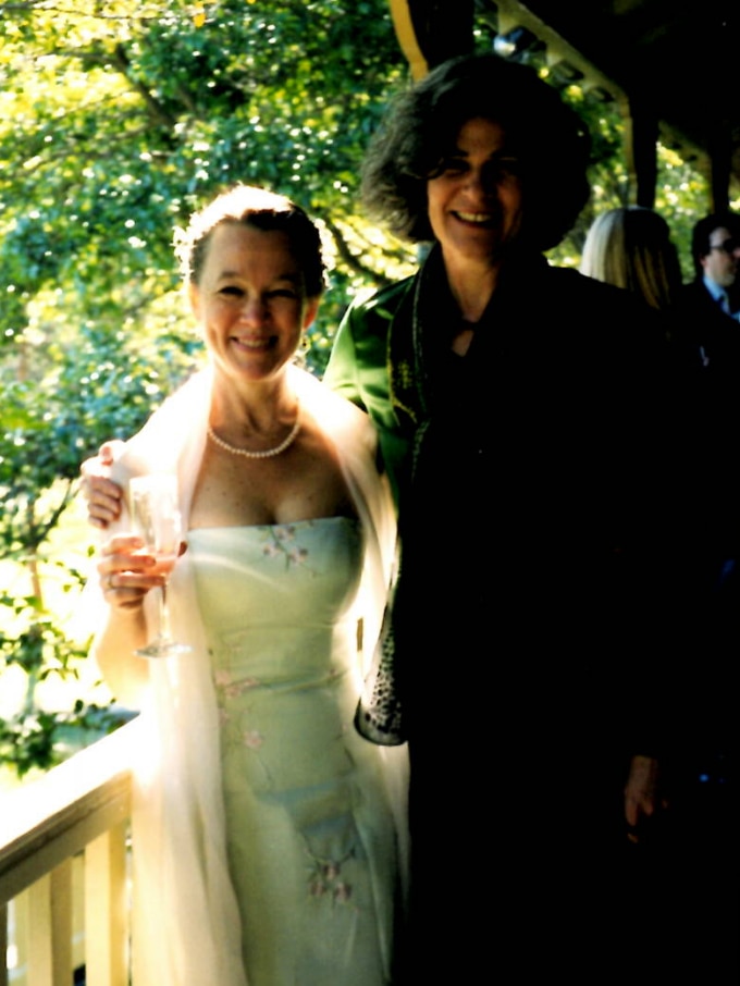 A woman wearing a white wedding dress, holding a glass of wine, standing with a taller woman with dark wavy hair wearing black.