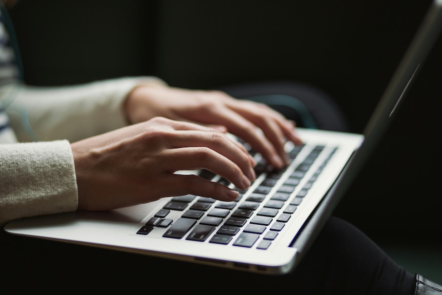 Close up shot of thin, white hands typing at a laptop, gray with black keys, dark background.