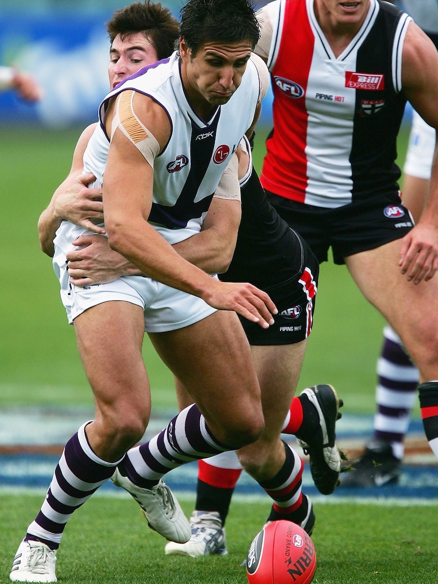 A Fremantle AFL player grimaces as he reaches for the ball while being grabbed from behind by a St Kilda defender.