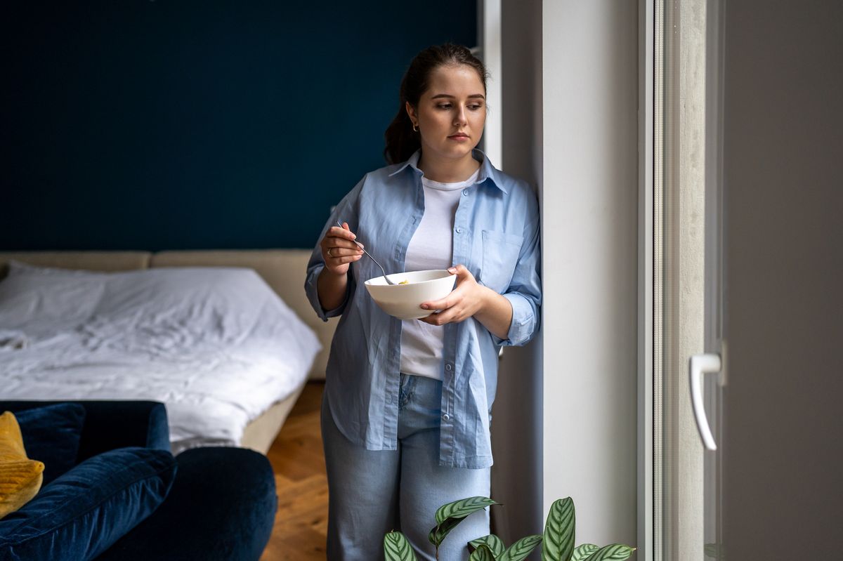 Woman standing by window with bowl of cereal, looking out window with upset face expression