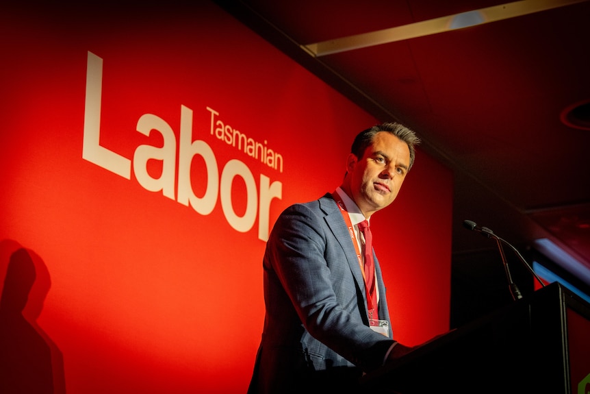 A man with short dark hair, wearing a blue suit and red tie, stands in front of a red wall.