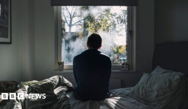 A man looks out of a window. He is in a dark room on an unmade bed. The atmosphere seems negative.