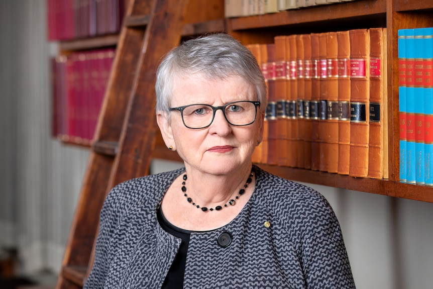 Virginia Bell has a serious expression and leans against a wooden bookshelf.
