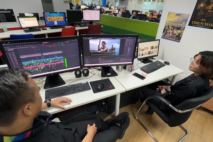 Two people sitting in front of the screens displaying a TV show scene.