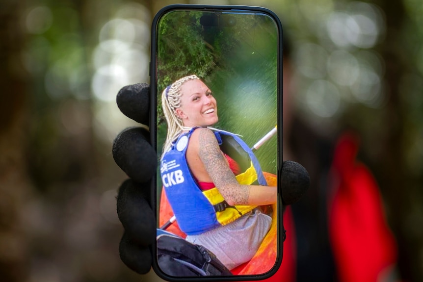 A woman wearing black and orange hi-vis is blurred in background, foreground she holds a phone showing image of young woman