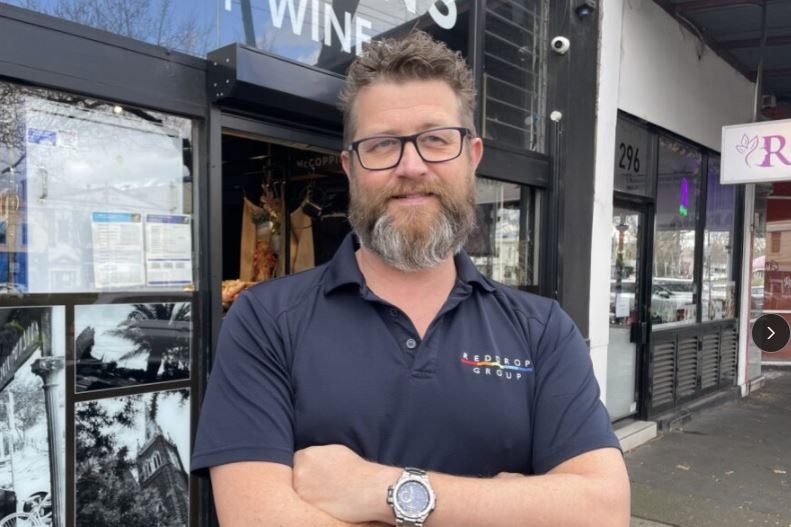 Lincoln smiles, has arms folded across chest. Caucasian, has brown hair and beard, glasses, stands in front of shop.