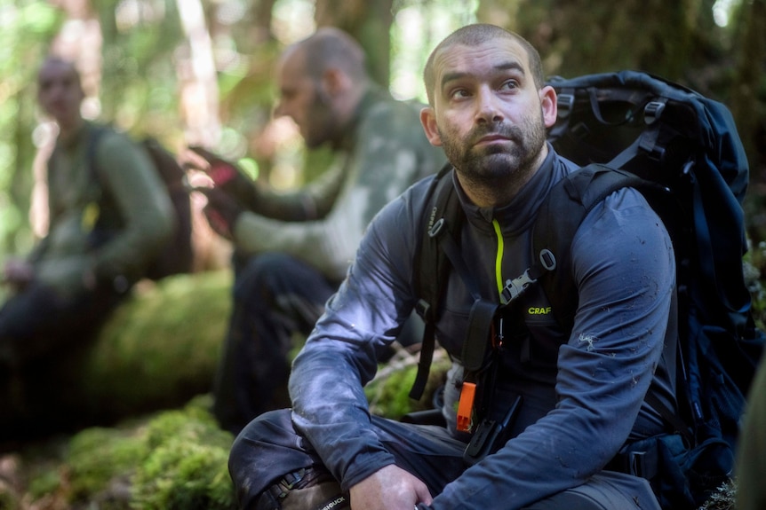 Man with a shaved hair cut wearing a backpack look upwards seated on a mossy tree root