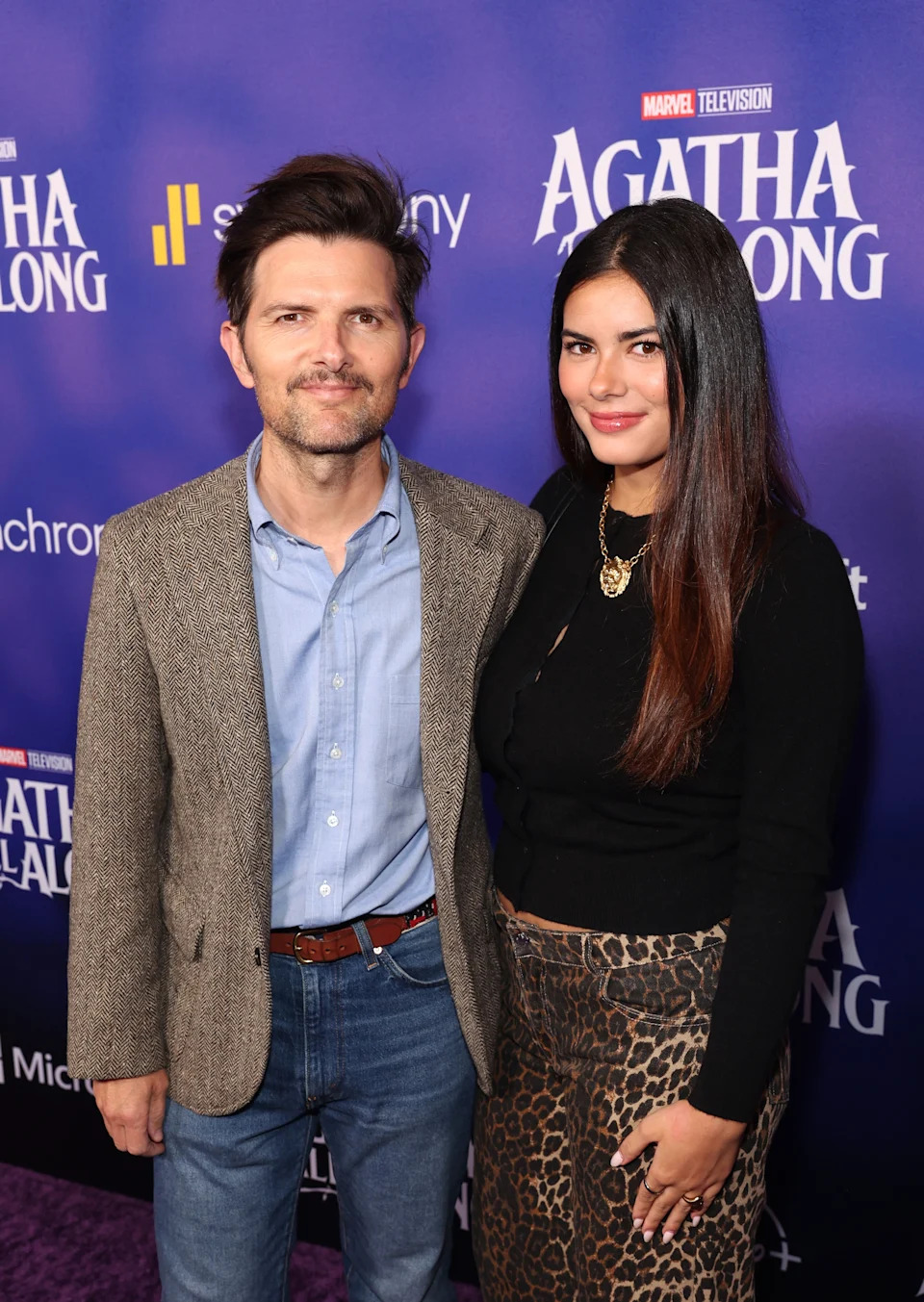 Adam Scott in a tweed blazer and jeans stands beside Frankie Scott in a top and leopard-print skirt at a media event