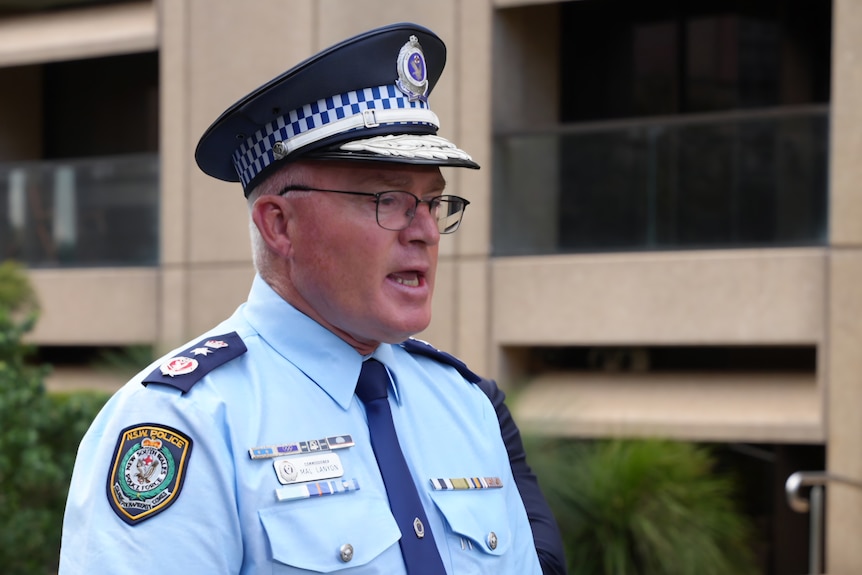 Mal Lanyon wears police uniform and hat and speaks to the media.