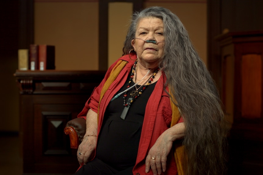 Vickie sits in a chair with her long hair falling over her shoulders as she looks at the camera