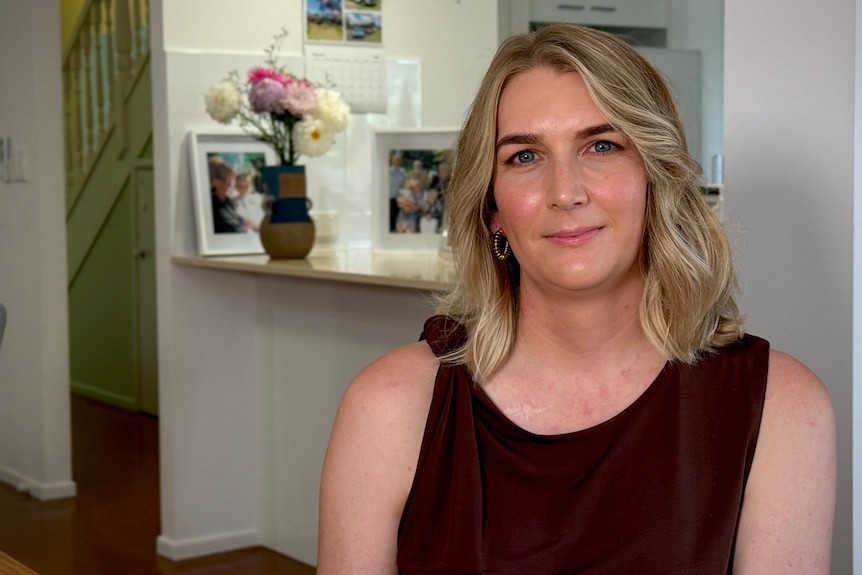 A woman with blonde shoulder length hair and a brown colour top looks at the camera