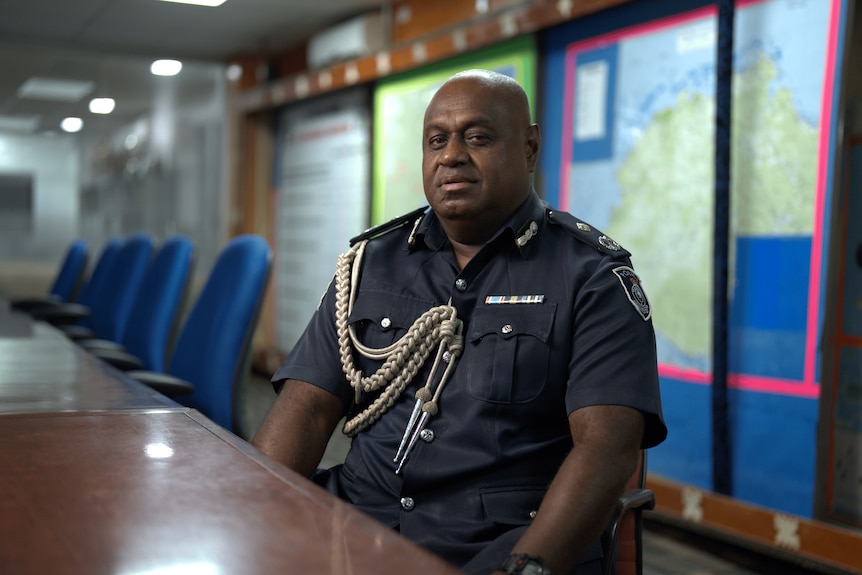 A man in a formal police uniform sits looking sternly at the camera in a room with a long empty table.