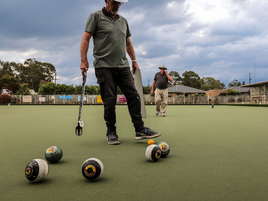 Bowls sit on the green surface of a lawn bowls rink with two men standing behind them.