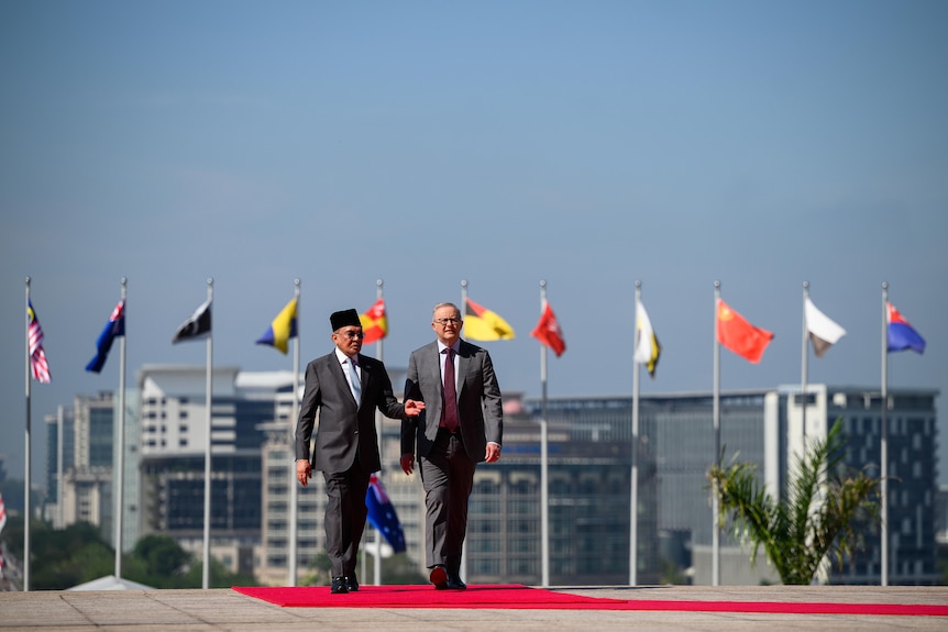 Two men in suit walk down a red carpet outdoors. A numvber of flags fly on poles behind them.