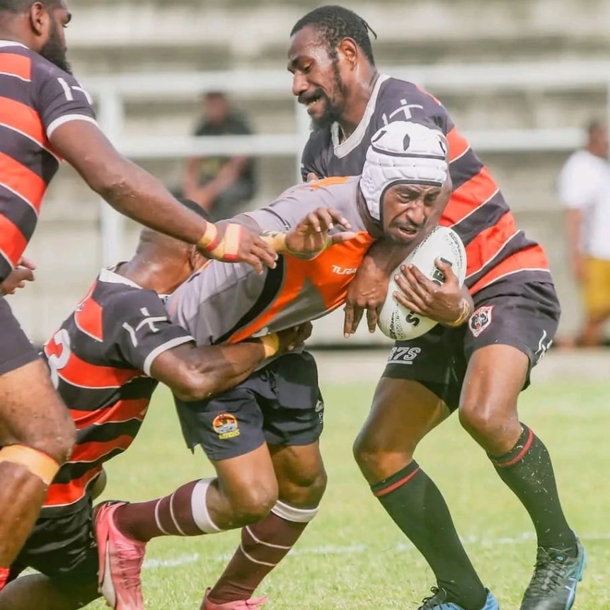 A man in white head gear wearing light grey and red league shirt breaks through a tackle while holding a white ball