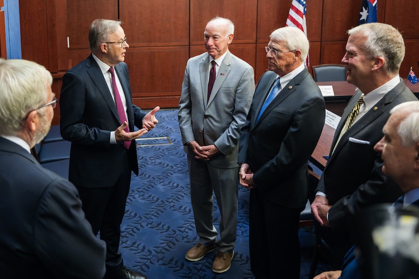A group of men stands in a room, speaking. There are US and Australia flags next to a table behind them.
