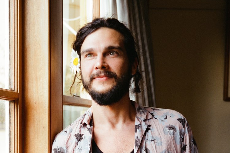 Luke Patterson, a First Nations man with a short beard, stands next to a window, wearing a black singlet and linen shirt.