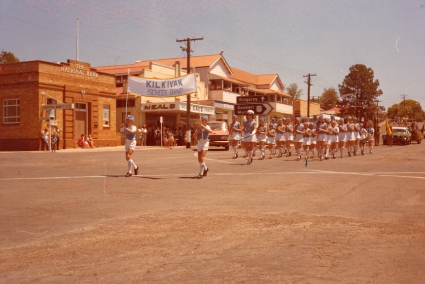 A group of children hold up a banner with their school's name on it as they march in a parade playing instruments.