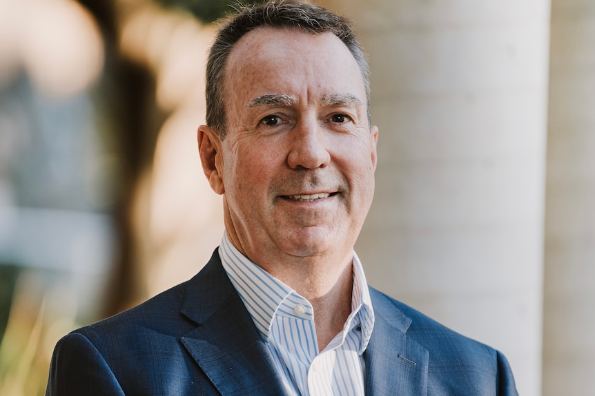 A smiling, middle aged man with short dark hair wears a suit while standing outdoors.