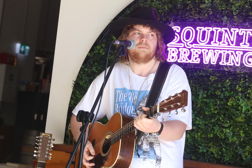 young man with red hair smiles at camera. He holds a guitar. There are speakers in the background. 